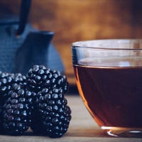 A glass cup filled with black raspberry tea sits on a wooden surface beside several blackberries, with a blurred dark teapot in the background.